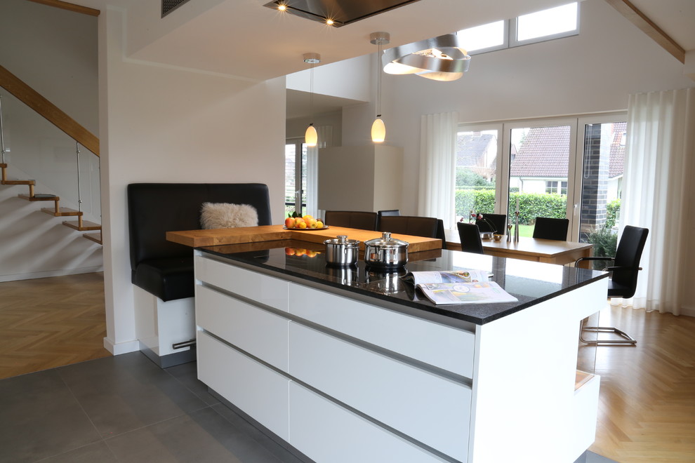 This is an example of a mid-sized contemporary galley eat-in kitchen in Bremen with flat-panel cabinets, white cabinets, with island, grey floor, black benchtop, a drop-in sink, granite benchtops, white splashback, glass sheet splashback, black appliances and cement tiles.
