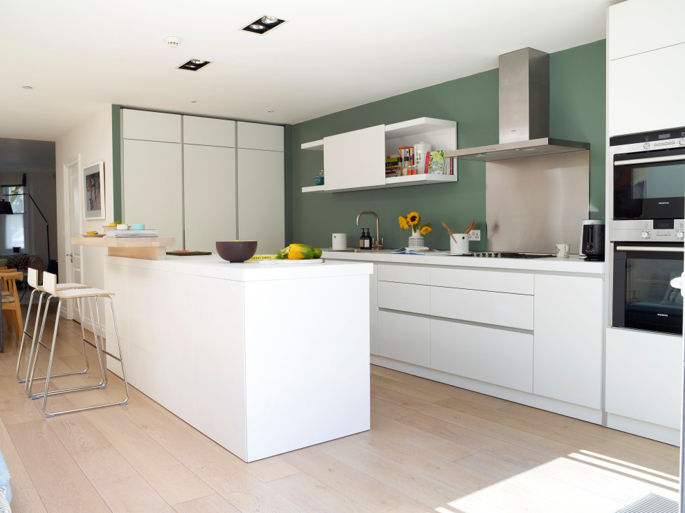 This is an example of a large contemporary u-shaped open plan kitchen with an integrated sink, flat-panel cabinets, white cabinets, laminate benchtops, metallic splashback, panelled appliances, with island, metal splashback, light hardwood floors, beige floor and white benchtop.