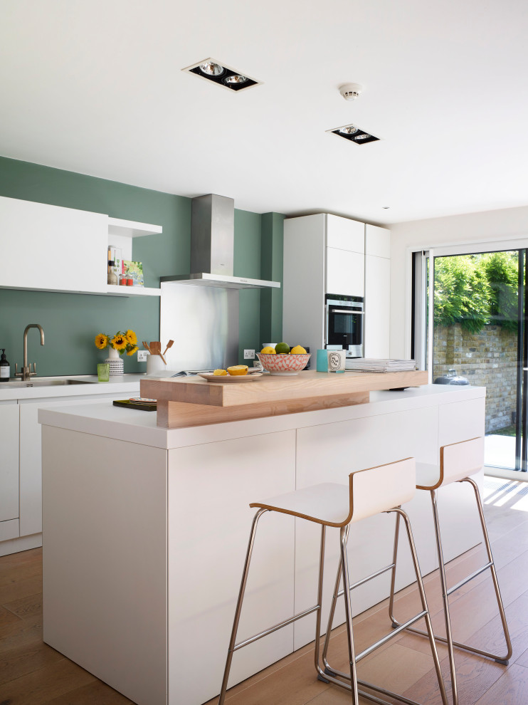 This is an example of a large contemporary u-shaped open plan kitchen in London with an integrated sink, flat-panel cabinets, white cabinets, laminate benchtops, green splashback, stainless steel appliances, light hardwood floors, with island, beige floor and white benchtop.