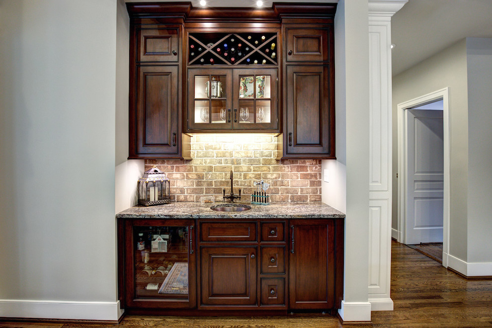 Photo of a small traditional single-wall wet bar in Other with an undermount sink, raised-panel cabinets, dark wood cabinets, granite benchtops, beige splashback, brick splashback, medium hardwood floors and brown floor.