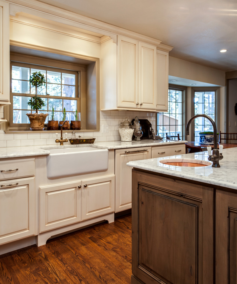 Bild på ett stort vit vitt kök, med en rustik diskho, luckor med upphöjd panel, vita skåp, marmorbänkskiva, vitt stänkskydd, stänkskydd i tunnelbanekakel, integrerade vitvaror, mellanmörkt trägolv, en köksö och brunt golv