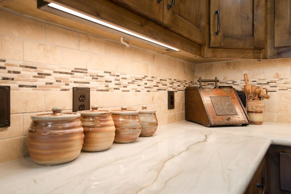 Eat-in kitchen - large transitional l-shaped brown floor and medium tone wood floor eat-in kitchen idea in Dallas with a farmhouse sink, recessed-panel cabinets, medium tone wood cabinets, multicolored backsplash, an island, quartzite countertops, travertine backsplash, stainless steel appliances and white countertops