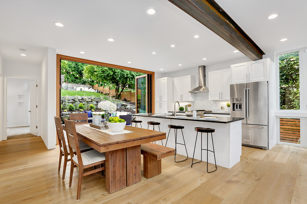 Photo of a mid-sized transitional galley eat-in kitchen in Seattle with white cabinets, quartzite benchtops, white splashback, ceramic splashback, stainless steel appliances, light hardwood floors, with island, black benchtop, shaker cabinets and beige floor.