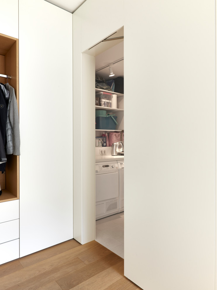 Photo of a large contemporary single-wall utility room in Munich with an integrated sink, flat-panel cabinets, white cabinets, concrete benchtops, white walls, medium hardwood floors, a side-by-side washer and dryer, brown floor and black benchtop.