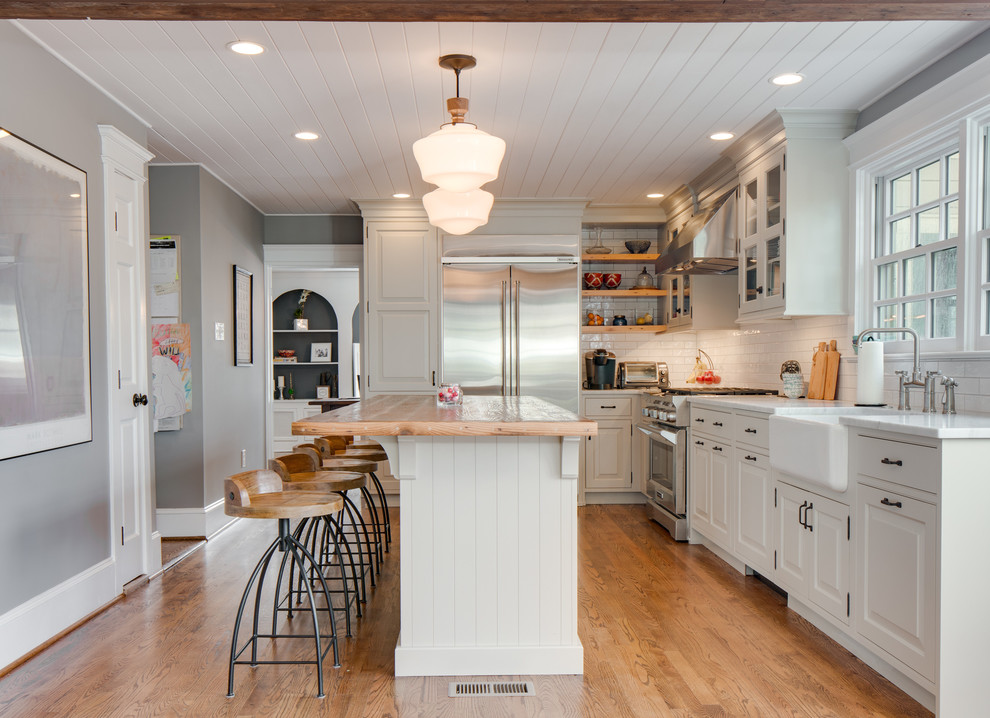 This is an example of a country l-shaped kitchen in Other with a farmhouse sink, raised-panel cabinets, white cabinets, white splashback, subway tile splashback, stainless steel appliances, medium hardwood floors, with island, brown floor and white benchtop.