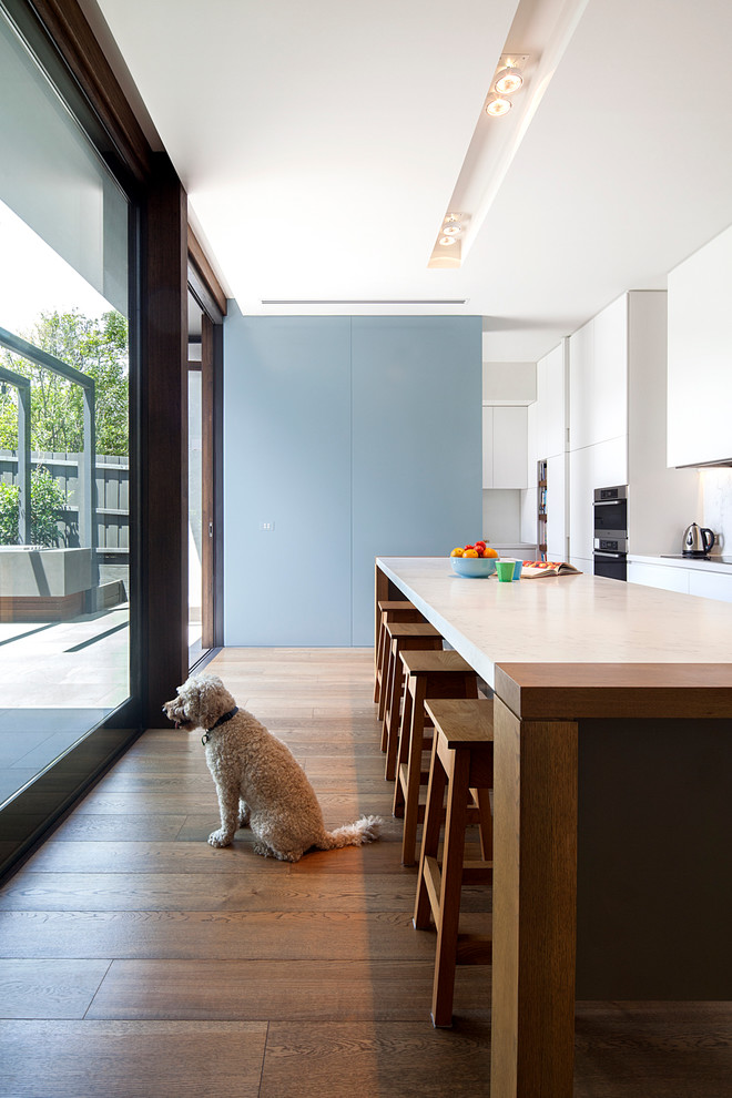This is an example of a mid-sized contemporary single-wall eat-in kitchen in Melbourne with a double-bowl sink, white cabinets, white splashback, stone slab splashback, stainless steel appliances, dark hardwood floors and with island.