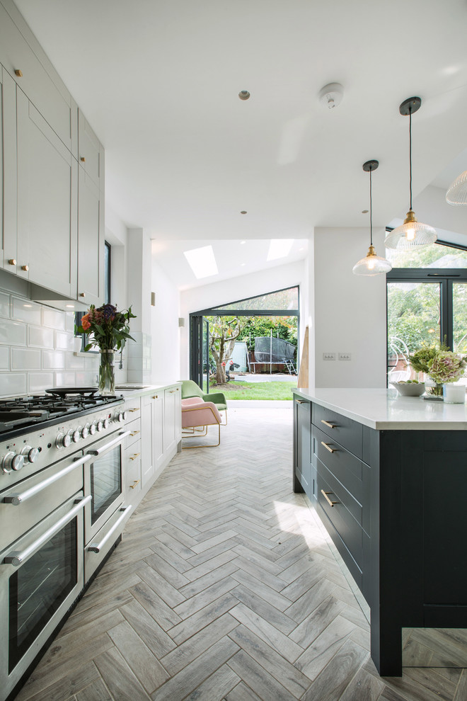 Transitional galley kitchen in London with shaker cabinets, grey cabinets, white splashback, stainless steel appliances, light hardwood floors, with island, grey floor and white benchtop.
