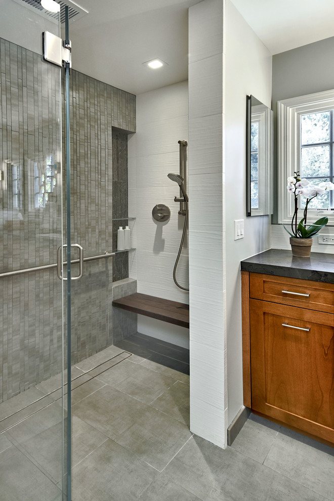 Photo of a mid-sized transitional master bathroom in San Francisco with shaker cabinets, medium wood cabinets, a curbless shower, a wall-mount toilet, gray tile, ceramic tile, grey walls, ceramic floors, granite benchtops, grey floor, a hinged shower door, black benchtops, a trough sink and a shower seat.