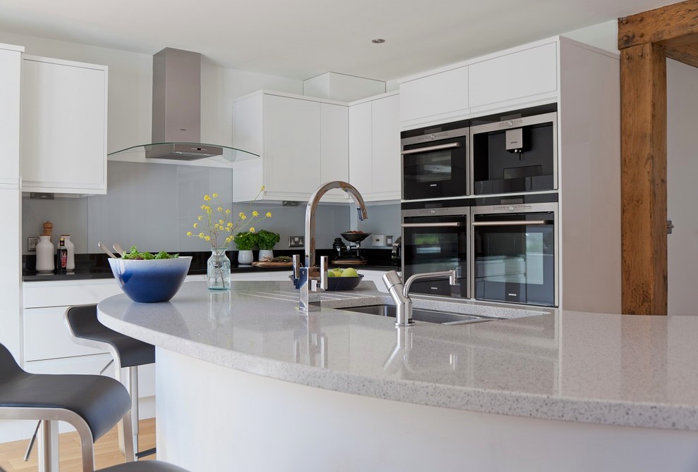 This is an example of a mid-sized contemporary l-shaped eat-in kitchen in Kent with an undermount sink, flat-panel cabinets, white cabinets, quartzite benchtops, grey splashback, glass sheet splashback, stainless steel appliances, light hardwood floors and with island.