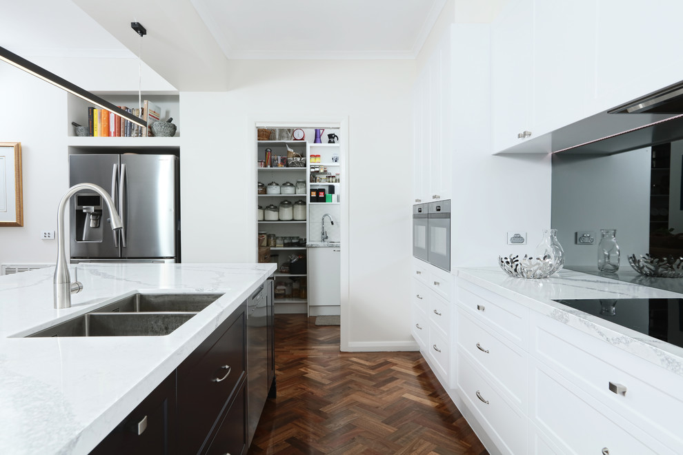 This is an example of a mid-sized transitional galley open plan kitchen in Melbourne with an undermount sink, shaker cabinets, quartz benchtops, mirror splashback, black appliances, dark hardwood floors and with island.