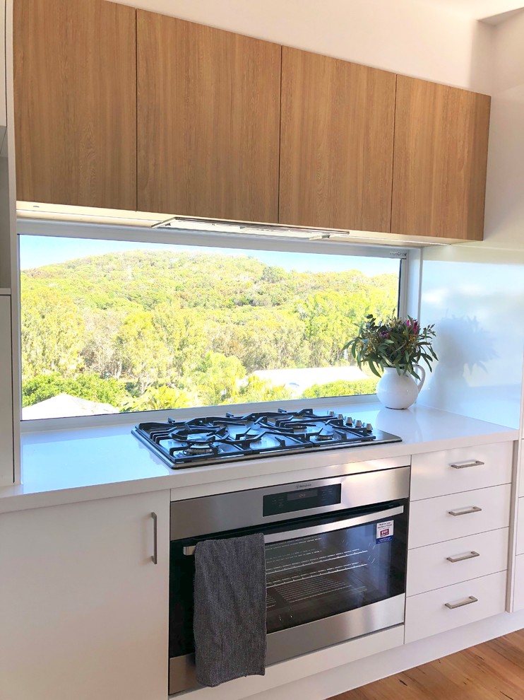 Photo of a mid-sized beach style l-shaped kitchen in Sunshine Coast with flat-panel cabinets, white cabinets, white splashback, window splashback, stainless steel appliances, light hardwood floors, with island and white benchtop.