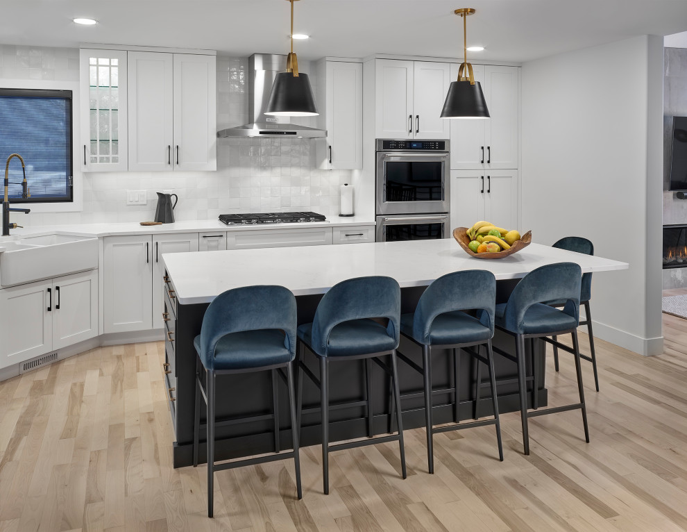 This is an example of a mid-sized country l-shaped eat-in kitchen in Edmonton with a farmhouse sink, shaker cabinets, white cabinets, quartz benchtops, white splashback, ceramic splashback, stainless steel appliances, light hardwood floors, with island, brown floor and white benchtop.