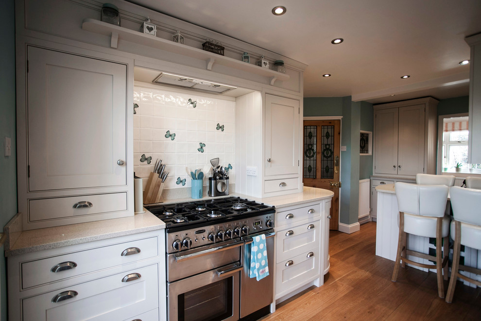 This is an example of a mid-sized country u-shaped eat-in kitchen in Kent with shaker cabinets, white cabinets, granite benchtops, medium hardwood floors, a single-bowl sink, white splashback, ceramic splashback, white appliances, with island and beige benchtop.