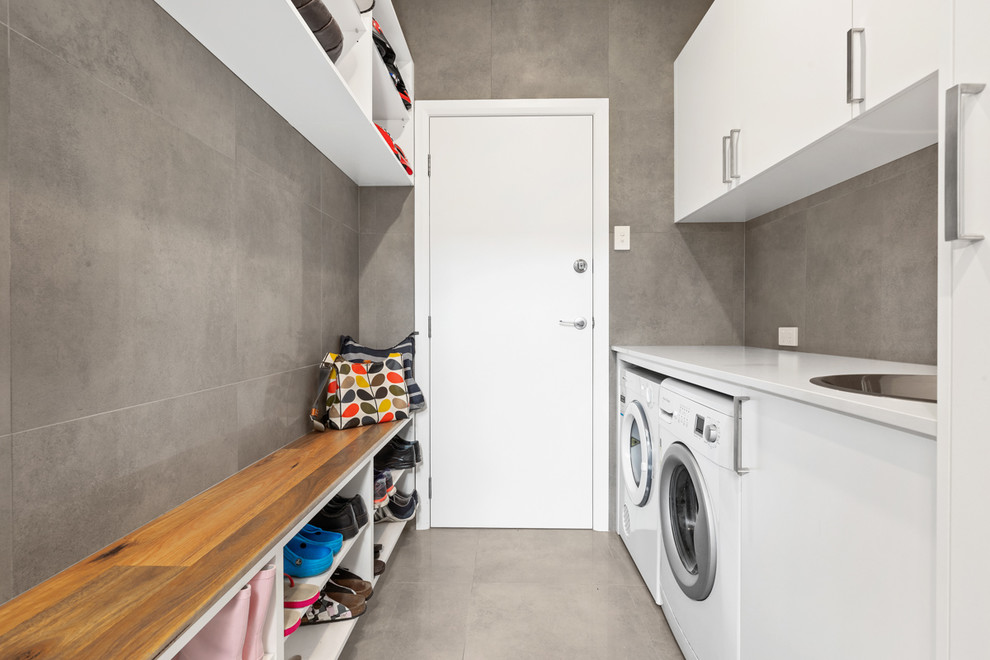 This is an example of a mid-sized contemporary galley utility room in Sydney with a drop-in sink, white cabinets, quartz benchtops, grey walls, porcelain floors, a side-by-side washer and dryer, grey floor, white benchtop, grey splashback and ceramic splashback.