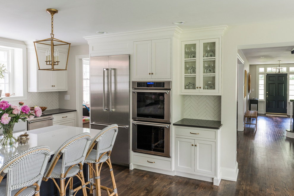 This is an example of a large transitional u-shaped kitchen pantry in Philadelphia with a farmhouse sink, shaker cabinets, white cabinets, quartz benchtops, white splashback, ceramic splashback, stainless steel appliances, dark hardwood floors, with island and brown floor.