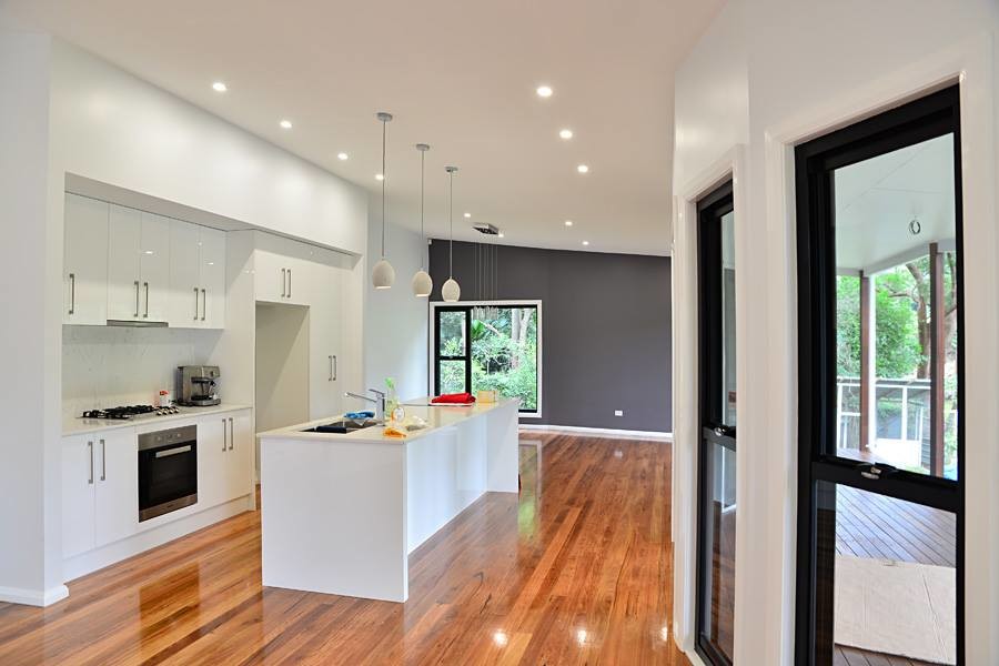 This is an example of a mid-sized beach style single-wall open plan kitchen in Wollongong with a double-bowl sink, flat-panel cabinets, white cabinets, quartz benchtops, white splashback, marble splashback, medium hardwood floors and with island.