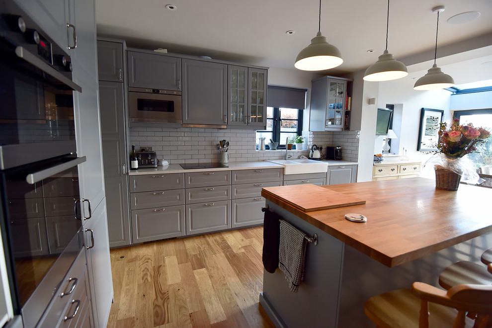 Photo of a large transitional l-shaped eat-in kitchen in Hertfordshire with a double-bowl sink, recessed-panel cabinets, grey cabinets, quartz benchtops, grey splashback, subway tile splashback, stainless steel appliances, medium hardwood floors and with island.