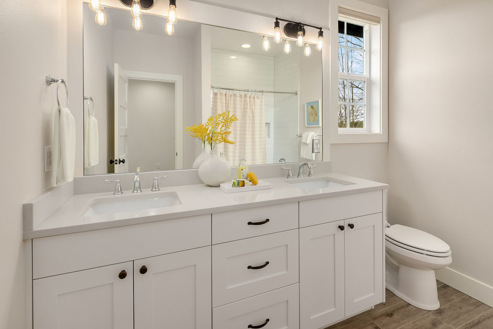 This is an example of a large transitional kids bathroom in Seattle with recessed-panel cabinets, white cabinets, a shower/bathtub combo, a one-piece toilet, white walls, medium hardwood floors, an undermount sink, brown floor, a shower curtain, white benchtops, a double vanity and a built-in vanity.