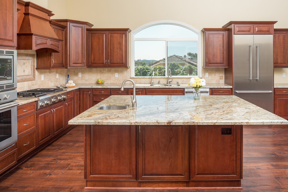 This is an example of an expansive traditional l-shaped open plan kitchen in San Diego with raised-panel cabinets, dark wood cabinets, granite benchtops, beige splashback, stone tile splashback, stainless steel appliances, dark hardwood floors, with island, an undermount sink, brown floor, beige benchtop and vaulted.