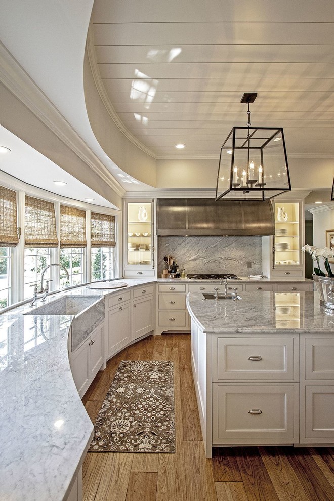 Example of a mid-sized transitional l-shaped light wood floor and brown floor enclosed kitchen design in Austin with a farmhouse sink, recessed-panel cabinets, white cabinets, quartzite countertops, gray backsplash, stone slab backsplash, stainless steel appliances and an island