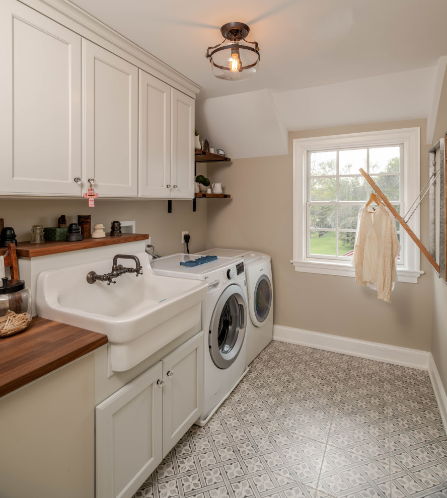 This is an example of a country single-wall dedicated laundry room in Philadelphia with a farmhouse sink, white cabinets, wood benchtops, ceramic floors, a side-by-side washer and dryer, grey floor and brown benchtop.