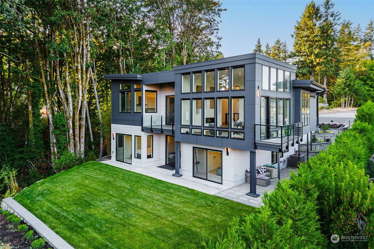 A modern home with large, floor-to-ceiling windows and a combination of white and dark grey siding. The spacious balcony overlooks a beautifully landscaped yard, perfect for outdoor entertainment.