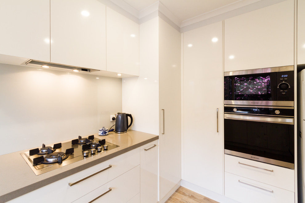 Photo of a mid-sized contemporary u-shaped eat-in kitchen in Melbourne with a drop-in sink, flat-panel cabinets, white cabinets, quartz benchtops, white splashback, glass sheet splashback, stainless steel appliances, light hardwood floors, a peninsula, brown floor and beige benchtop.