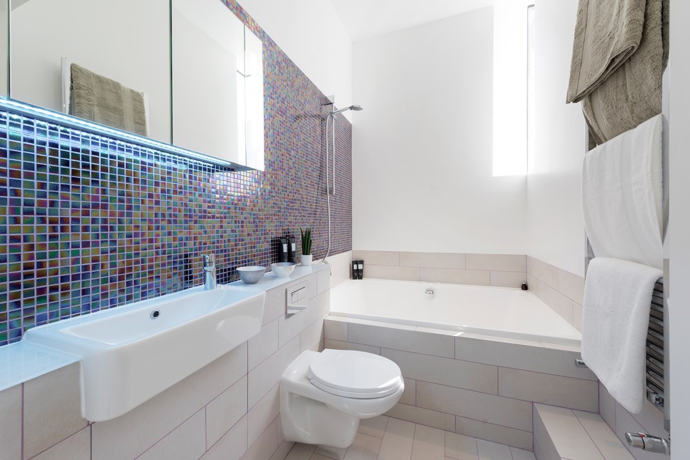 Mid-sized contemporary master bathroom in London with a drop-in tub, a shower/bathtub combo, a wall-mount toilet, mosaic tile, white walls, multi-coloured tile, a wall-mount sink and beige floor.