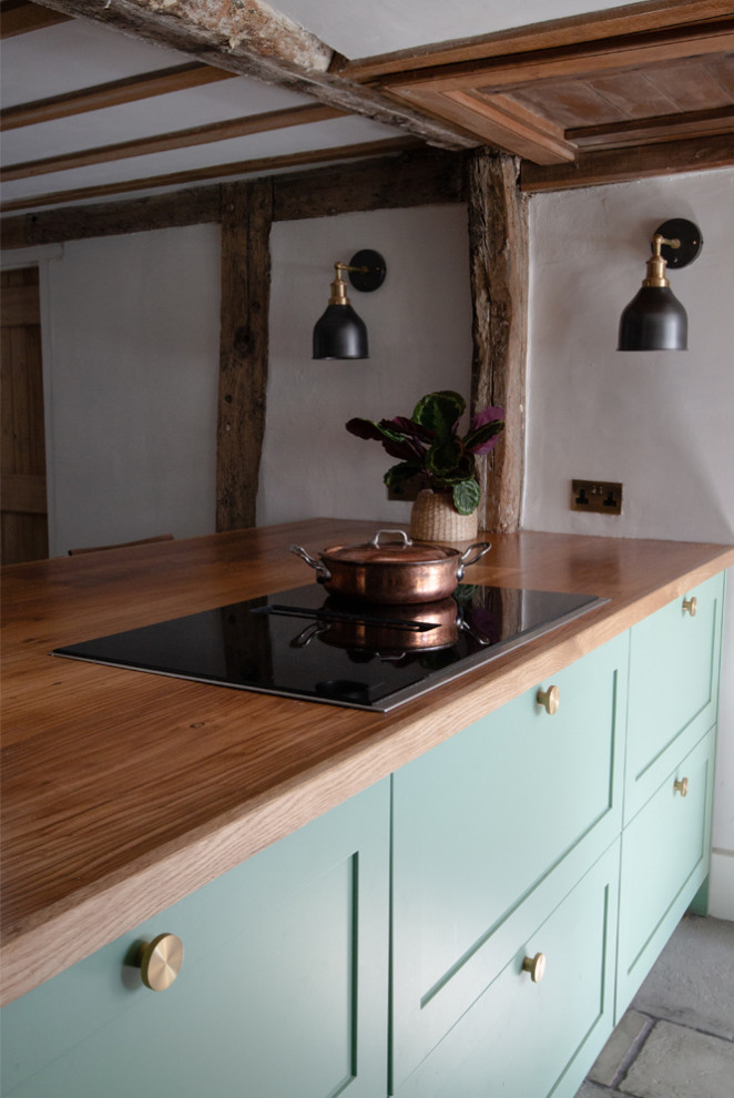 Medium sized contemporary u-shaped kitchen/diner in Oxfordshire with a belfast sink, shaker cabinets, green cabinets, wood worktops, stainless steel appliances, limestone flooring, an island, grey floors, brown worktops and exposed beams.
