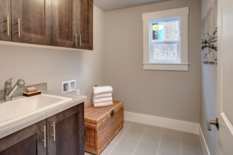 This is an example of a mid-sized traditional single-wall dedicated laundry room in Seattle with a drop-in sink, flat-panel cabinets, dark wood cabinets, tile benchtops, grey walls, ceramic floors and a side-by-side washer and dryer.