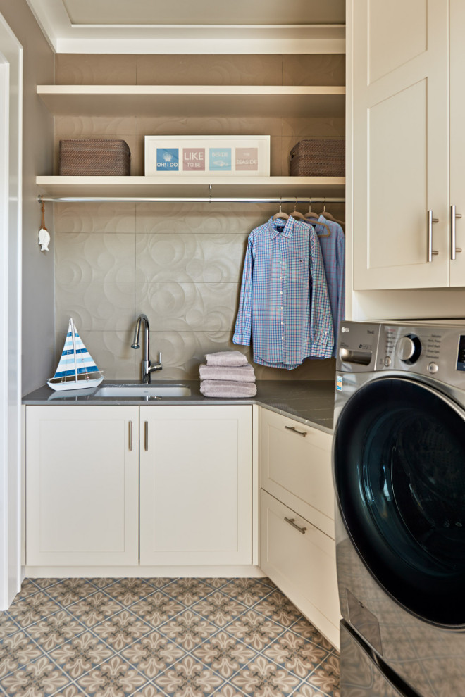 Photo of a mid-sized beach style l-shaped dedicated laundry room in Wilmington with an undermount sink, shaker cabinets, beige cabinets, marble benchtops, beige splashback, porcelain splashback, grey walls, ceramic floors, a side-by-side washer and dryer, multi-coloured floor and grey benchtop.
