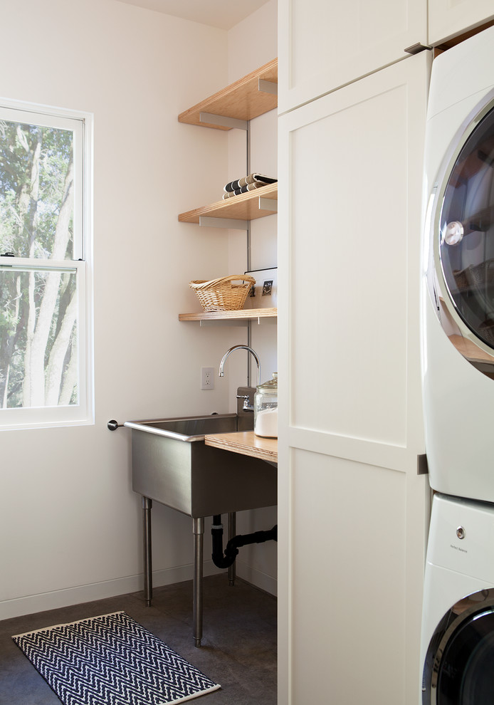 Mid-sized country dedicated laundry room in San Francisco with an utility sink, white cabinets, white walls, concrete floors, a stacked washer and dryer, shaker cabinets, wood benchtops, grey floor and beige benchtop.
