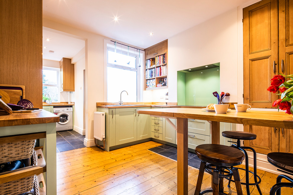 Inspiration for a small country eat-in kitchen in Other with a double-bowl sink, shaker cabinets, green cabinets, wood benchtops, green splashback, glass sheet splashback, stainless steel appliances, light hardwood floors, a peninsula, brown floor and brown benchtop.