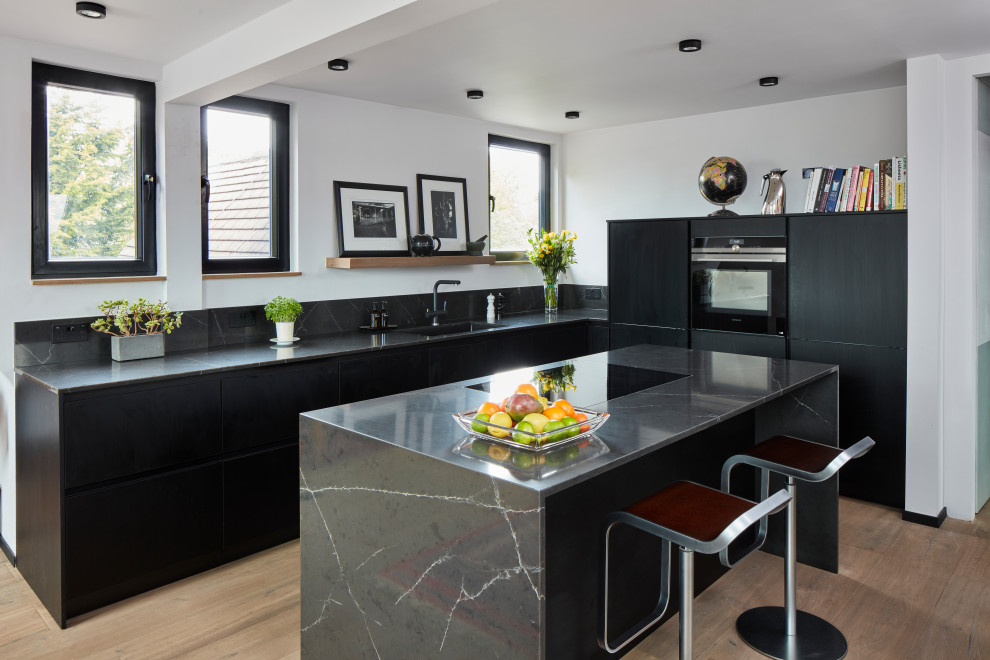 Photo of a small contemporary grey and black l-shaped open plan kitchen in London with a submerged sink, flat-panel cabinets, black cabinets, quartz worktops, grey splashback, engineered quartz splashback, stainless steel appliances, light hardwood flooring, an island, brown floors and grey worktops.