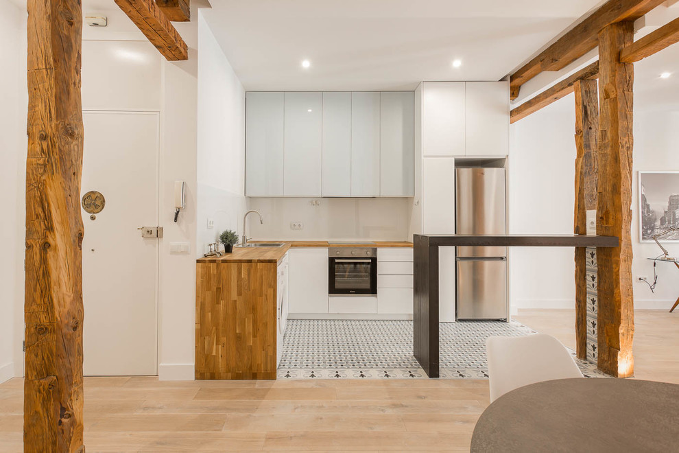 Design ideas for a mid-sized scandinavian l-shaped open plan kitchen in Madrid with flat-panel cabinets, wood benchtops, white splashback, glass sheet splashback, stainless steel appliances, ceramic floors, a peninsula, multi-coloured floor, a drop-in sink and blue cabinets.