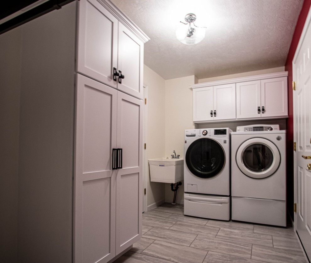 Modelo de cuarto de lavado tradicional renovado pequeño con pila para lavar, armarios con paneles lisos, puertas de armario blancas, suelo de baldosas de cerámica, lavadora y secadora juntas y suelo gris