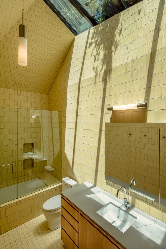 На фото: ванная комната среднего размера с плоскими фасадами, светлыми деревянными фасадами, полновстраиваемой ванной, душем над ванной, унитазом-моноблоком, желтой плиткой, керамической плиткой, желтыми стенами, полом из керамической плитки, врезной раковиной, мраморной столешницей, желтым полом, душем с распашными дверями, белой столешницей, тумбой под одну раковину и встроенной тумбой