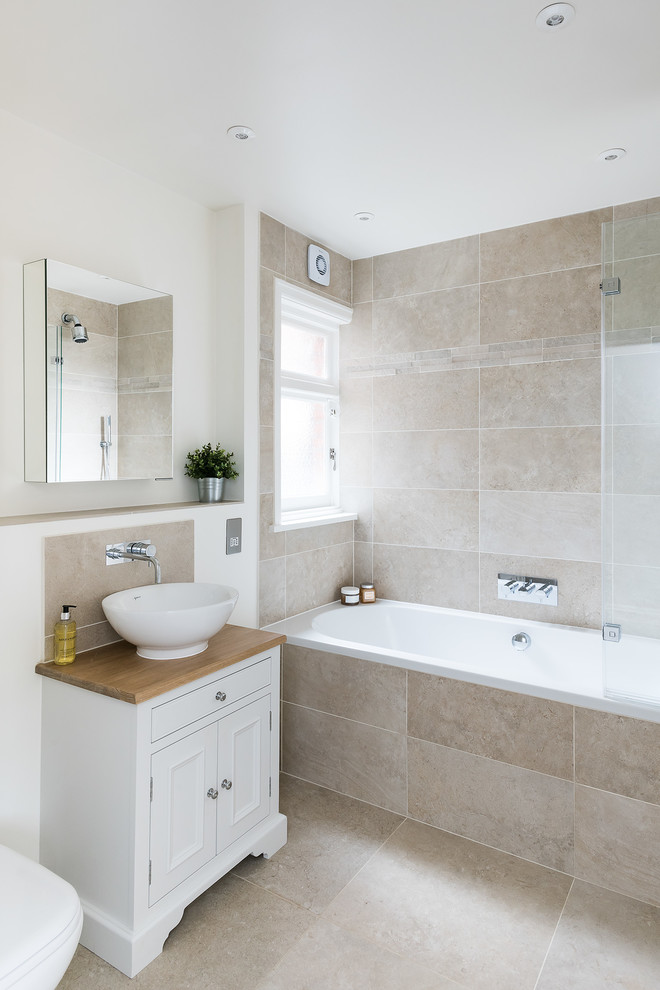 Mid-sized traditional kids bathroom in London with furniture-like cabinets, white cabinets, a drop-in tub, a shower/bathtub combo, a two-piece toilet, beige tile, travertine, beige walls, travertine floors, a vessel sink, wood benchtops, beige floor and a hinged shower door.