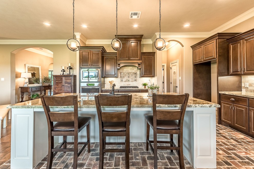 Example of a mid-sized mountain style u-shaped brick floor and brown floor open concept kitchen design in Austin with a farmhouse sink, shaker cabinets, medium tone wood cabinets, granite countertops, beige backsplash, travertine backsplash, stainless steel appliances and an island