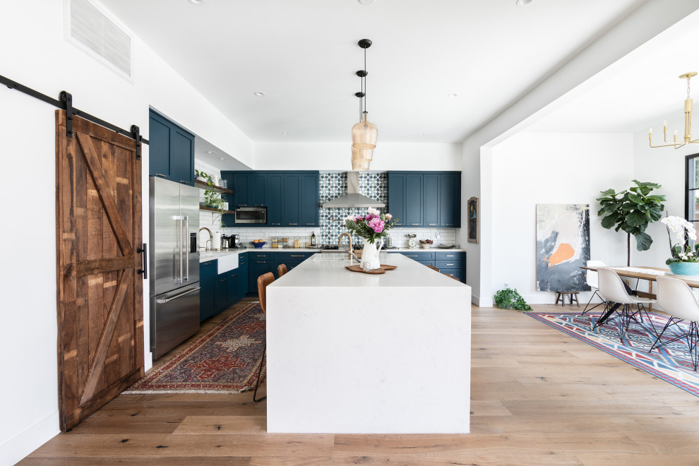 Mid-sized mediterranean l-shaped eat-in kitchen in Los Angeles with a farmhouse sink, shaker cabinets, blue cabinets, quartz benchtops, white splashback, ceramic splashback, stainless steel appliances, medium hardwood floors, with island, brown floor and white benchtop.