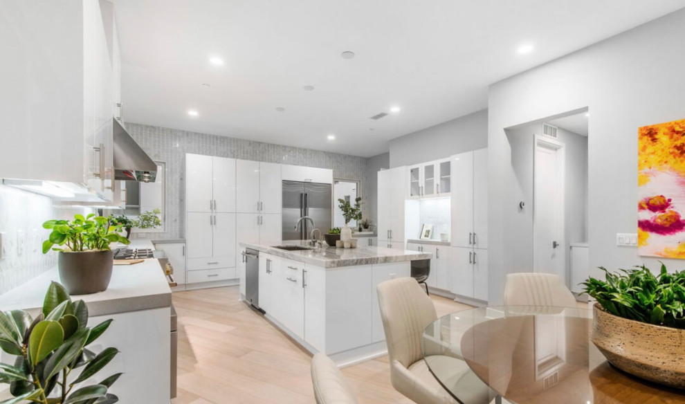 This is an example of a medium sized traditional u-shaped open plan kitchen in Los Angeles with a single-bowl sink, flat-panel cabinets, white cabinets, marble worktops, grey splashback, marble splashback, stainless steel appliances, light hardwood flooring, an island and grey worktops.