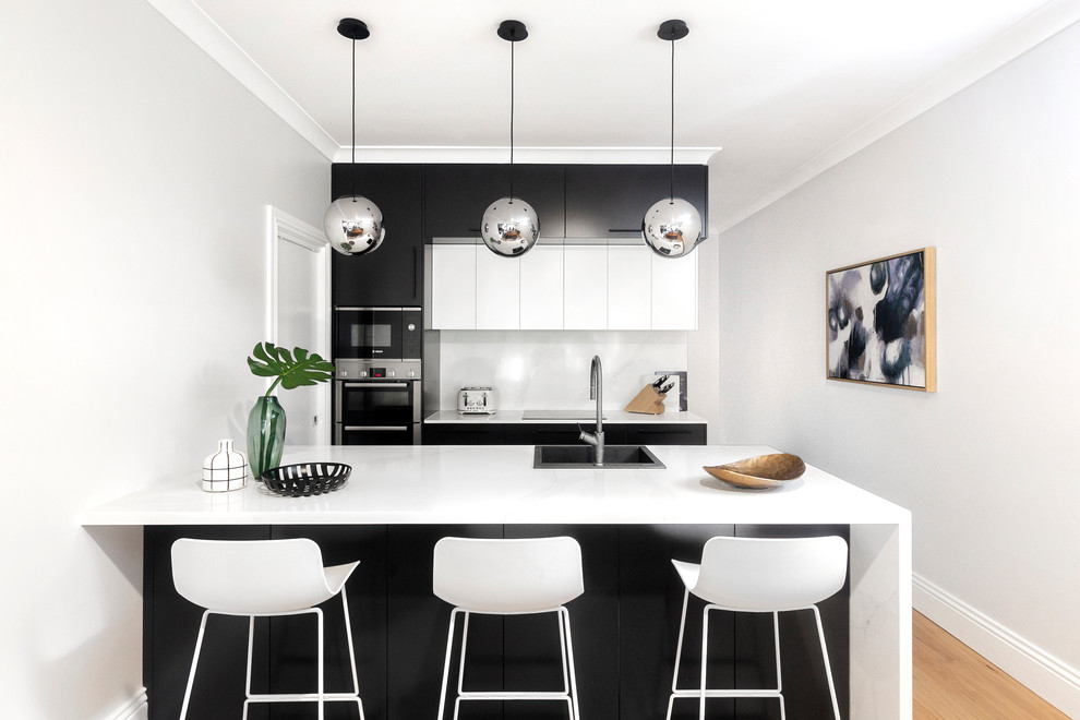 Design ideas for a contemporary galley kitchen in Sydney with a drop-in sink, flat-panel cabinets, black cabinets, white splashback, stainless steel appliances, medium hardwood floors, a peninsula, brown floor and white benchtop.