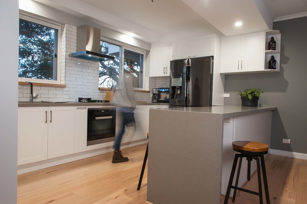 Mid-sized scandinavian u-shaped open plan kitchen in Melbourne with shaker cabinets, white cabinets, solid surface benchtops, with island, an undermount sink, white splashback, subway tile splashback, stainless steel appliances and light hardwood floors.