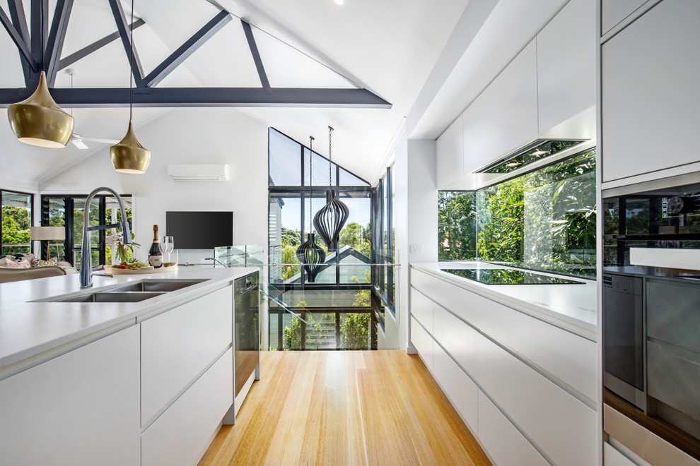 Mid-sized beach style l-shaped open plan kitchen in Sunshine Coast with an undermount sink, flat-panel cabinets, white cabinets, quartz benchtops, window splashback, black appliances, light hardwood floors, with island, multi-coloured floor and white benchtop.