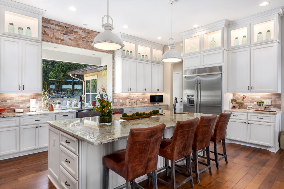 Photo of a transitional u-shaped kitchen in Austin with a farmhouse sink, recessed-panel cabinets, white cabinets, granite benchtops, multi-coloured splashback, stone tile splashback, stainless steel appliances, medium hardwood floors, with island, brown floor and beige benchtop.