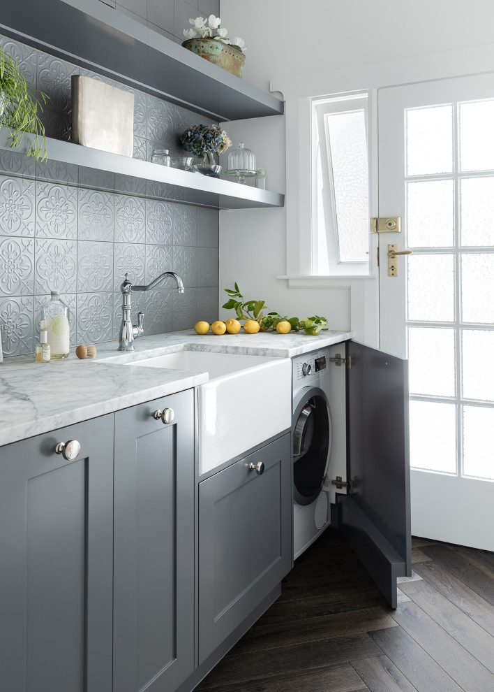 Photo of a mid-sized country single-wall dedicated laundry room in Auckland with a farmhouse sink, shaker cabinets, grey cabinets, quartzite benchtops, grey splashback, ceramic splashback, white walls, dark hardwood floors, a concealed washer and dryer, black floor and white benchtop.