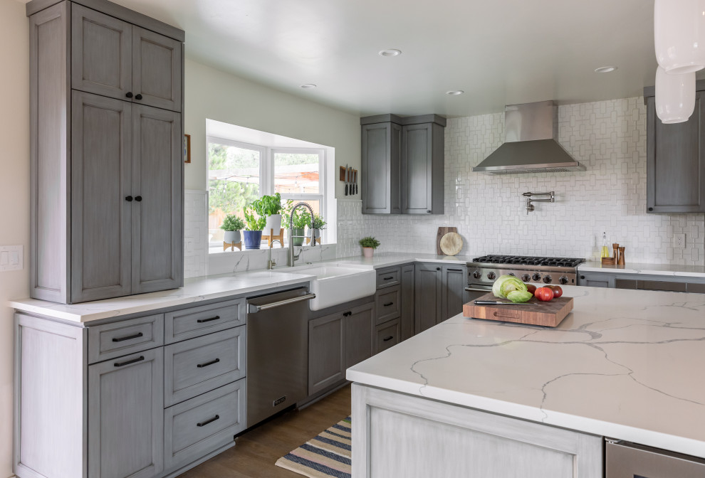 This is an example of a medium sized retro kitchen/diner in San Diego with a belfast sink, shaker cabinets, grey cabinets, engineered stone countertops, white splashback, ceramic splashback, stainless steel appliances, vinyl flooring, an island, brown floors and white worktops.