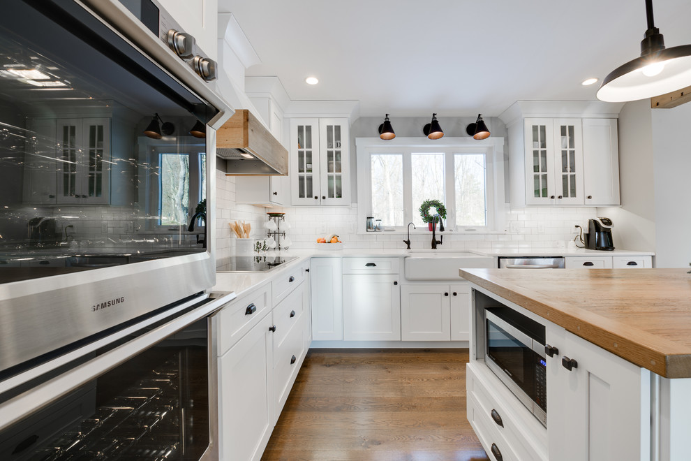 Design ideas for a country l-shaped kitchen in Philadelphia with a farmhouse sink, shaker cabinets, white cabinets, white splashback, subway tile splashback, stainless steel appliances, medium hardwood floors, with island, brown floor and white benchtop.