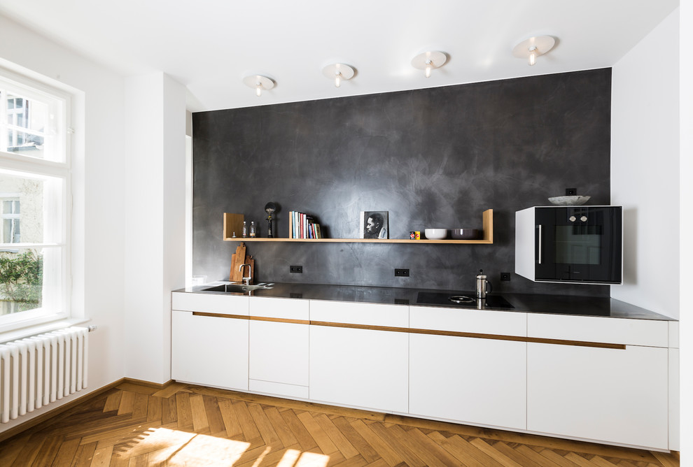 This is an example of a large modern single-wall open plan kitchen in Munich with an integrated sink, flat-panel cabinets, white cabinets, stainless steel benchtops, black splashback, limestone splashback, black appliances, medium hardwood floors, no island and brown floor.
