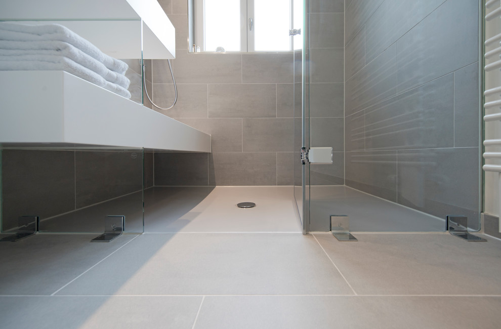 Small contemporary 3/4 bathroom in Stuttgart with flat-panel cabinets, white cabinets, a curbless shower, gray tile, ceramic tile, grey walls, ceramic floors, a trough sink, solid surface benchtops, grey floor and a hinged shower door.